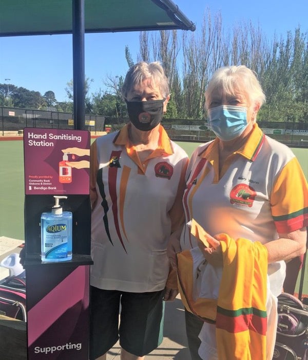 Photo of two women standing beside hand sanitising station at bowling club